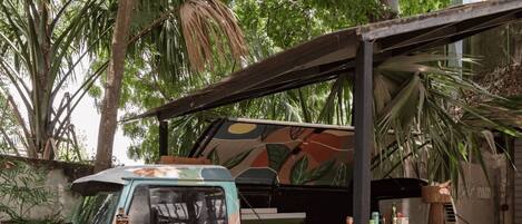 A food truck with a colorful design parked under a shelter, surrounded by green chairs and a table.