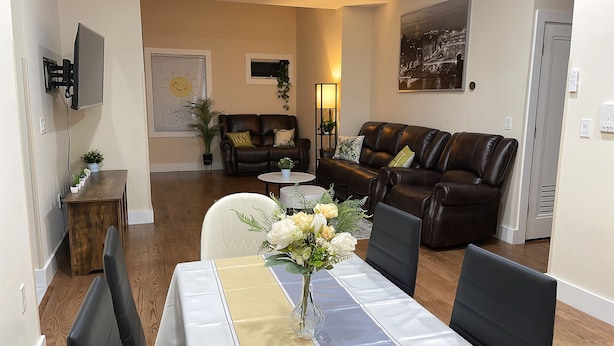 A living room with a dining table, two sofas, a TV mounted on the wall, and a cityscape painting.