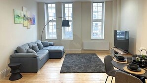 A modern living room with a grey sectional sofa, a black floor lamp, a flat-screen TV, and a dining area with a table and chairs. - Spacious flat in the East End

 (London)