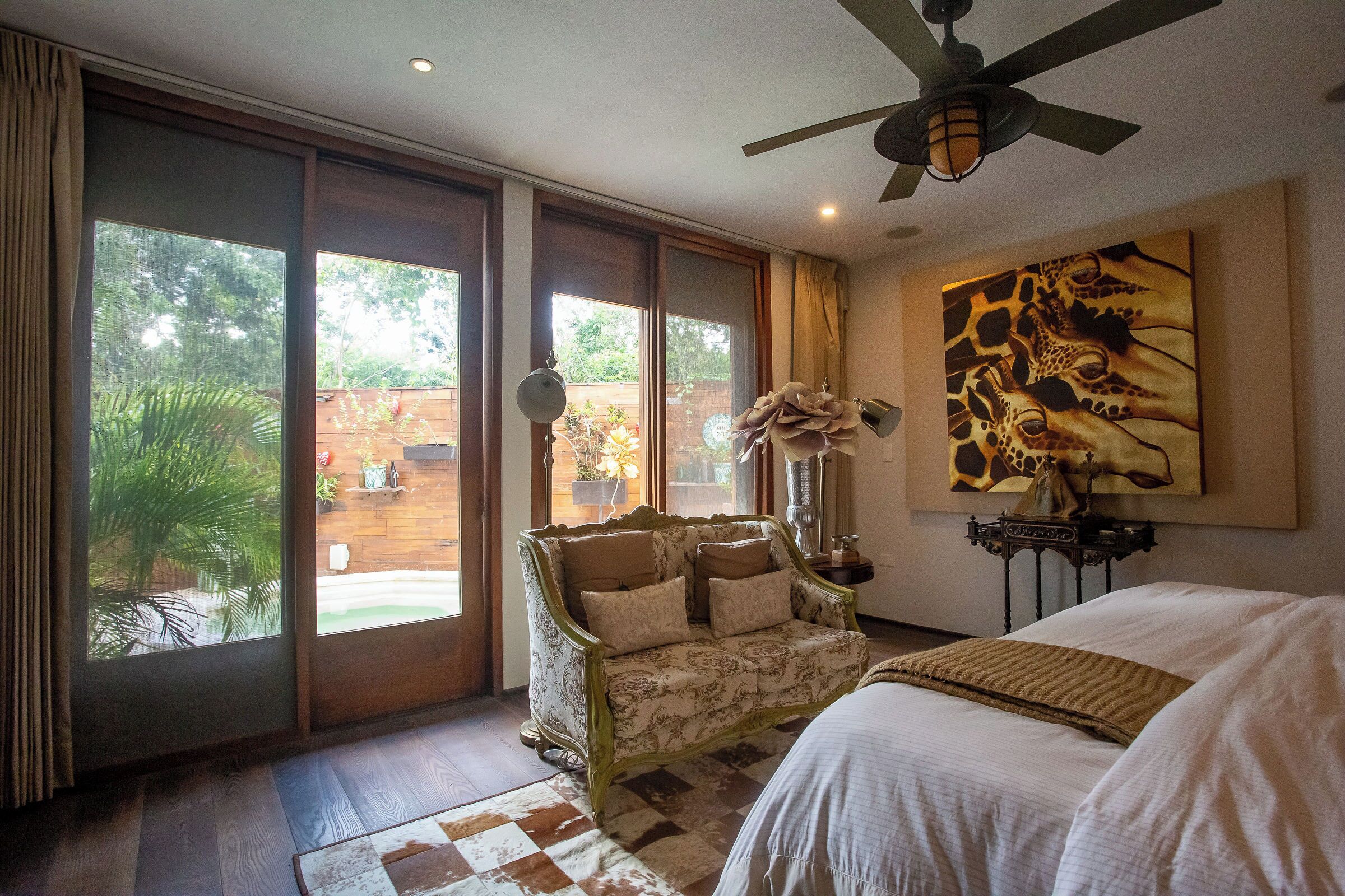 A bedroom with a large bed, a chaise lounge, a ceiling fan, and a view of the outdoors.