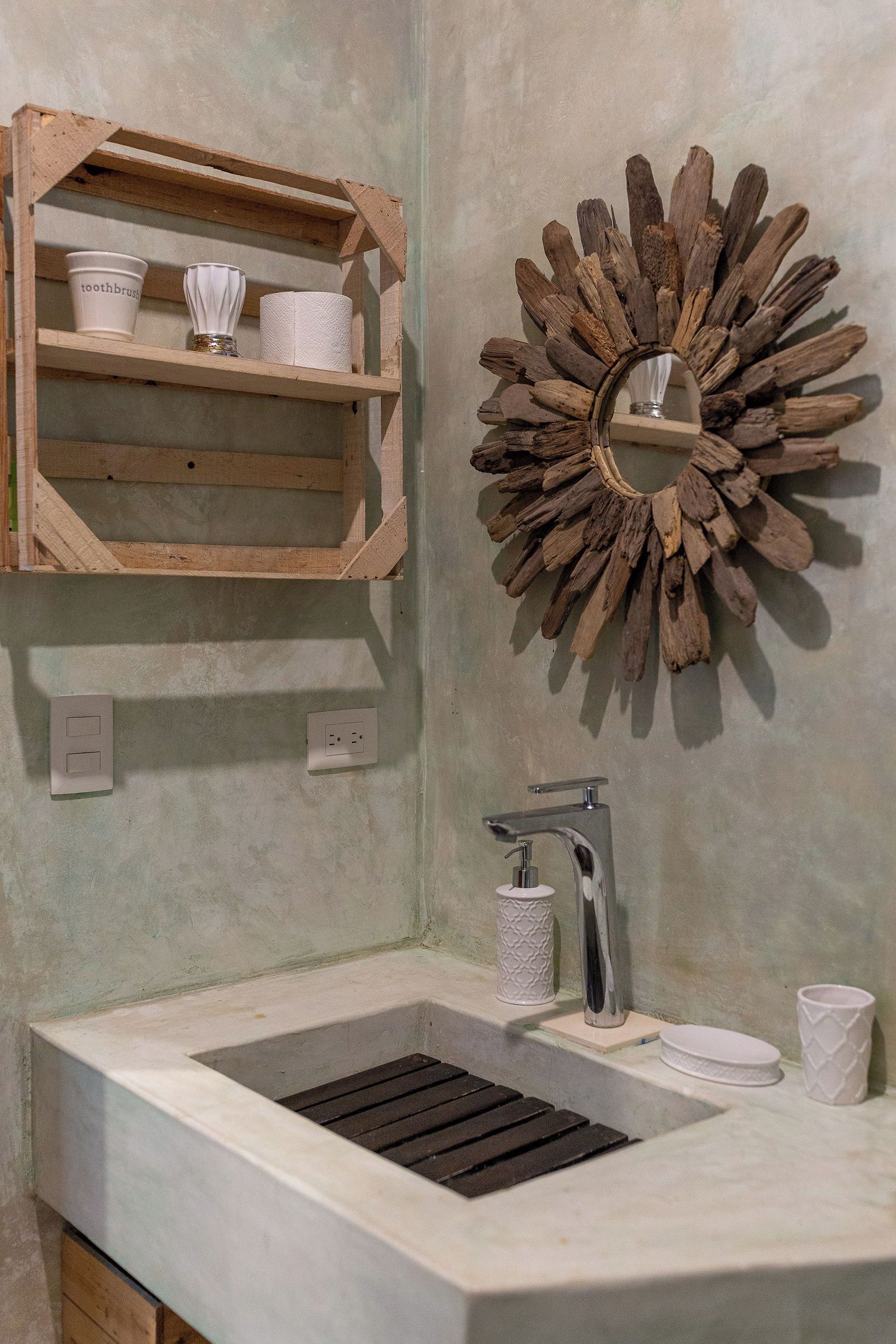 Un baño con estante de madera que contiene artículos de tocador, un adorno de pared rústico, un lavabo con grifo moderno y un espejo pequeño.