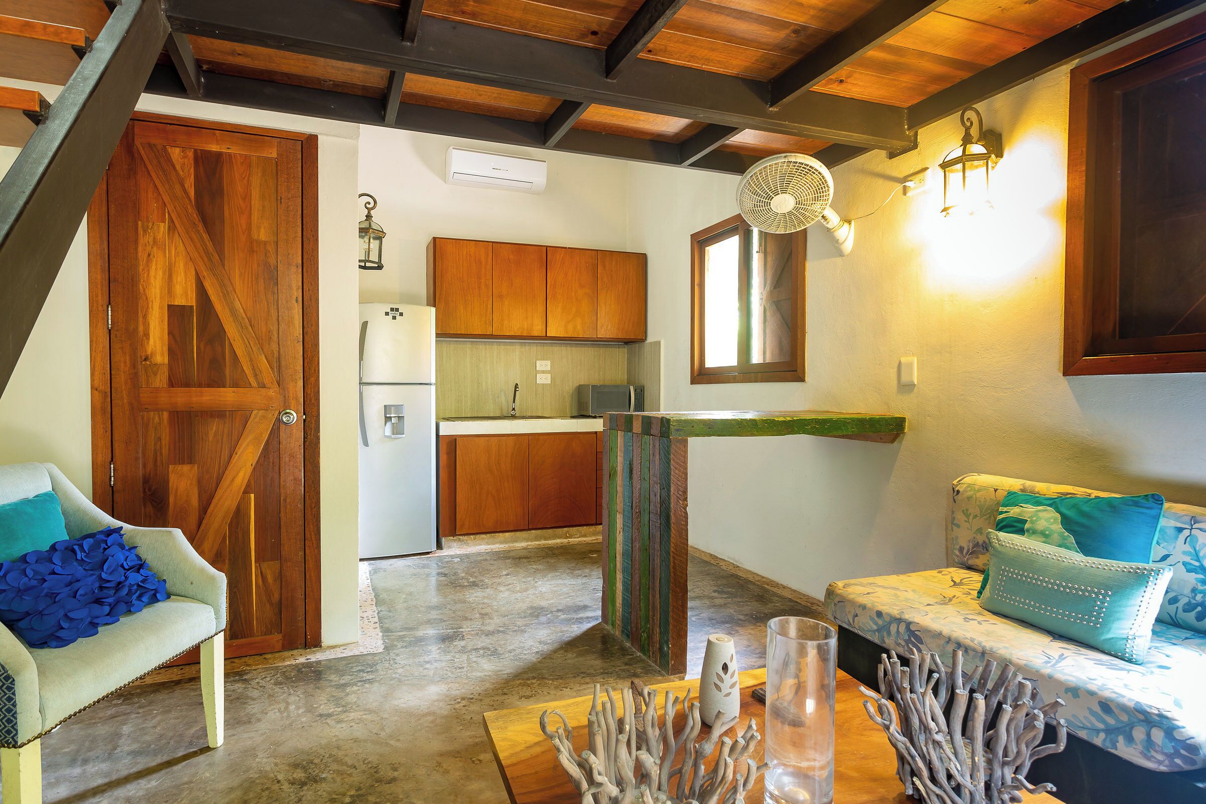 A cozy living area with a wooden staircase, a kitchen with wooden cabinets, a sofa with patterned cushions, and a decorative plant centerpiece.