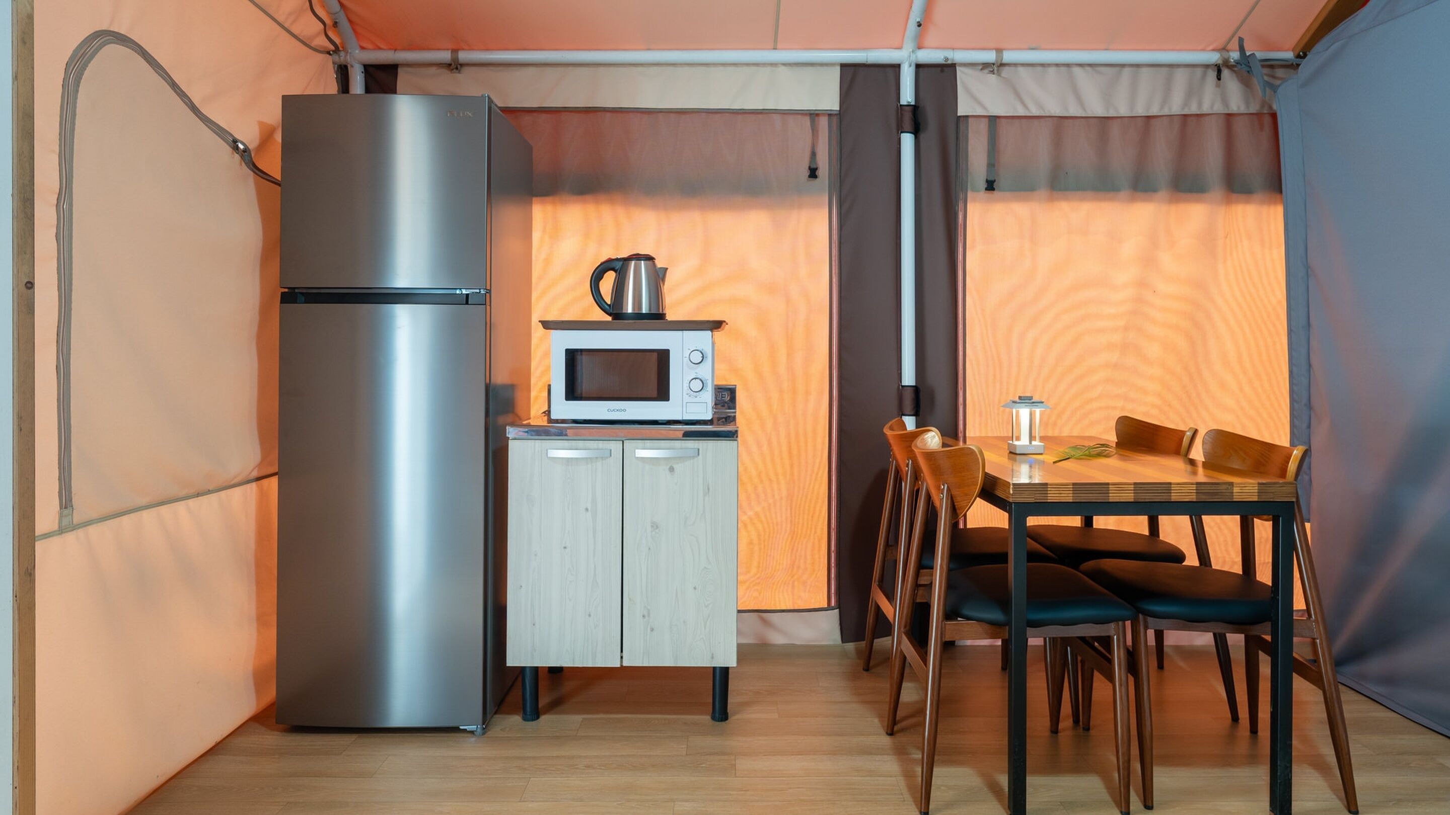 A compact kitchen with a refrigerator, microwave, and kettle, a wooden cabinet, and a dining table with chairs.