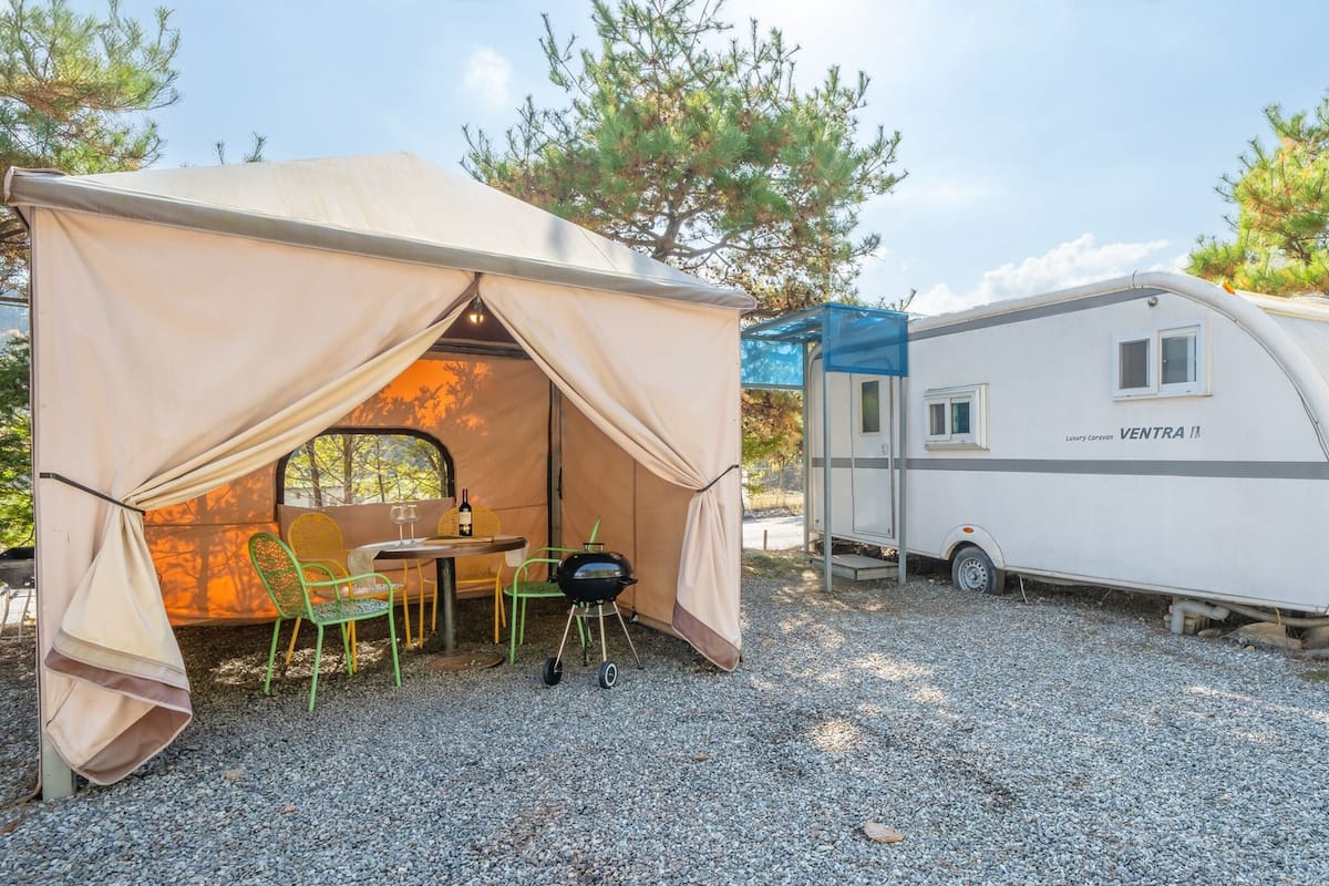 Une tente avec une table et des chaises, un barbecue et une caravane de luxe dotée d’une fenêtre.