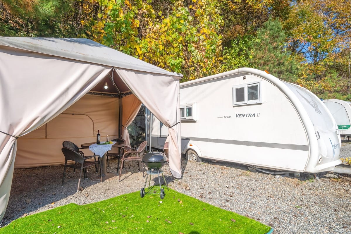 Une caravane de luxe avec une tente attenante, comprenant un coin repas avec chaises et table, un barbecue et une petite fenêtre.