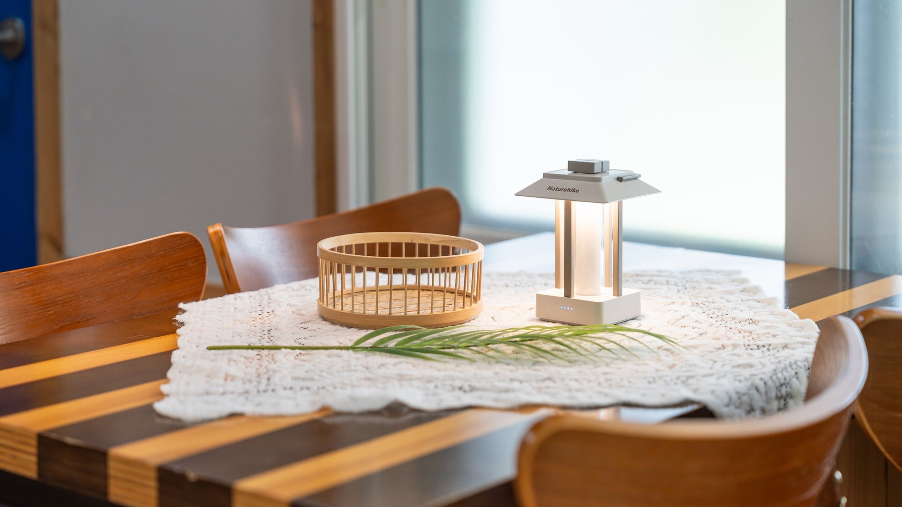 A dining table with a white lace tablecloth, a wooden chair, a small wooden basket, and a white table lamp with a label.
