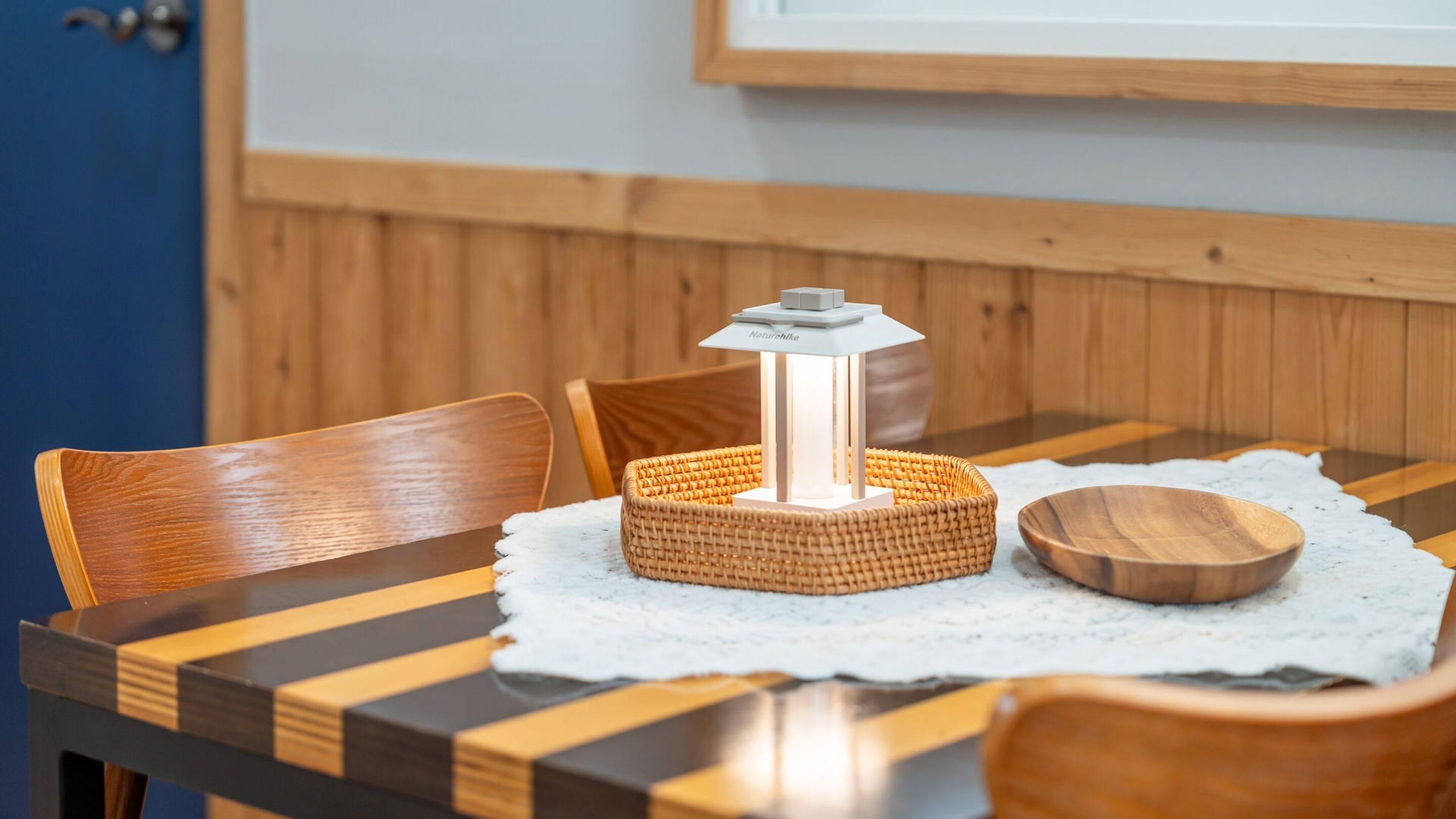A dining table with a woven placemat, a wooden bowl, and a table lamp.