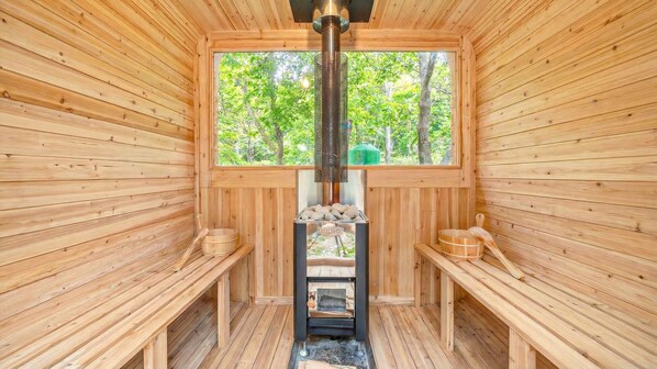 A sauna room with wooden walls, a central heating unit, benches, and a window with a view of trees.