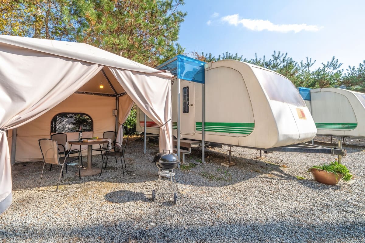 Un terrain de camping avec une tente installée pour servir de salle à manger, un barbecue et une caravane garée sur un sol de gravier.