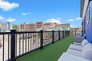 En balkon med kunstgræs, en hvid sofa og udsigt til bygninger og parkerede biler.