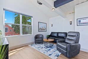 A modern living room with a black leather sofa, two armchairs, a round coffee table, and a large window offering a view of a red building and trees.