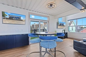 A modern interior with a round table and blue cushioned chairs, large windows offering views of buildings, and a ceiling fan.