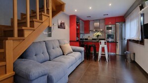 Ein modernes Wohnzimmer mit grauem Sofa, einer Holztreppe und einer Küche mit roten Schränken und Edelstahlgeräten.