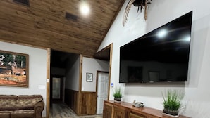 A spacious living area with a wooden ceiling, a large TV mounted on the wall, a wooden cabinet, and a patterned sofa.