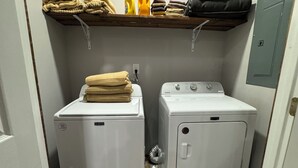 A laundry room with two stacked washer and dryer units, a shelf above holding folded towels and cleaning supplies, and a green bottle of detergent on the floor.