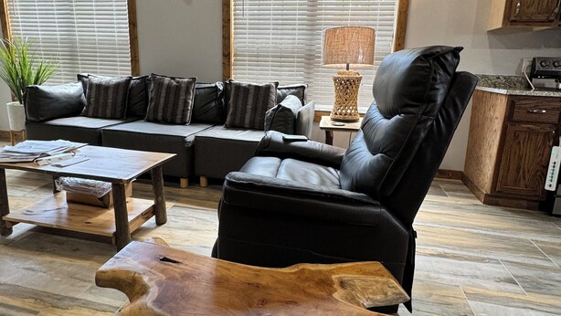 A rustic living room with a leather sofa, a wooden coffee table, and a dining table. There are two windows with blinds, a lamp, and a kitchen area with cabinets.