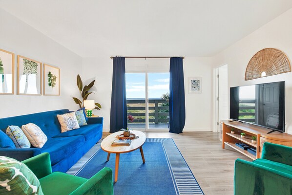 Een woonkamer met een blauwe bank, groene fauteuils, een salontafel, een televisie en een groot raam met gordijnen.