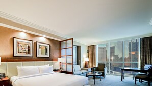 Une chambre d’hôtel avec un grand lit, deux tables de chevet avec des lampes, un bureau avec une chaise, un canapé et une grande fenêtre donnant sur la ville.