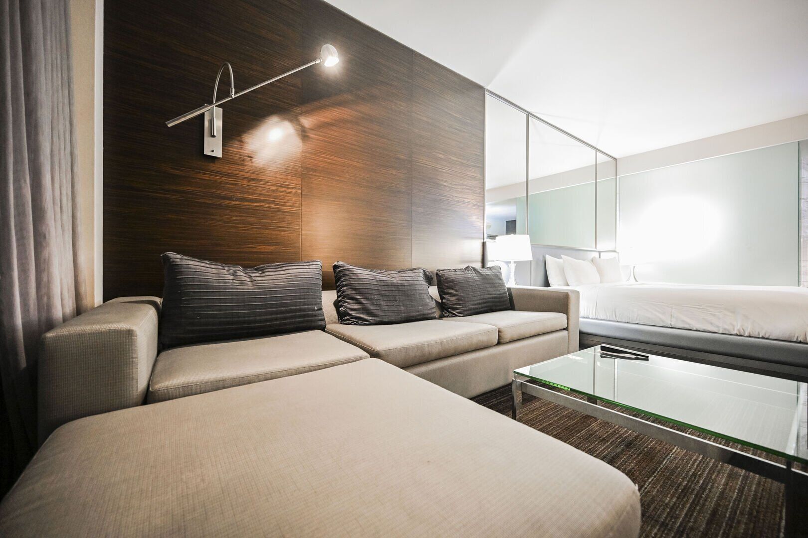 A modern hotel room with a sofa, a glass coffee table, a bed, and a wall-mounted lamp.