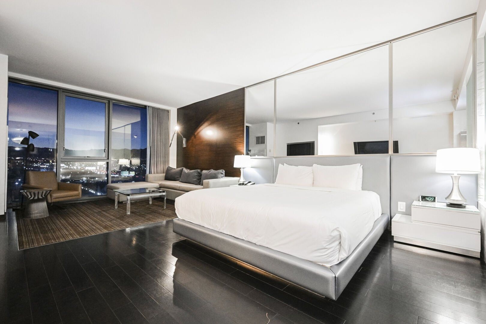 Une chambre d’hôtel moderne dotée d’un grand lit, d’un coin salon avec un canapé et des fauteuils, d’une table basse, d’une grande fenêtre donnant sur la ville et d’un miroir au mur.