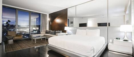 Une chambre d’hôtel moderne dotée d’un grand lit, d’un coin salon avec un canapé et des fauteuils, d’une table basse, d’une grande fenêtre donnant sur la ville et d’un miroir au mur.