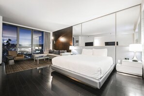 A modern hotel room with a large bed, a seating area with a sofa and armchairs, a coffee table, a large window with a city view, and a mirror on the wall.