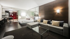 A modern hotel room with a large bed, a sofa, a glass coffee table, and a kitchen area visible through a glass partition. - SVS: High Floor Strip View Studio | Open Balcony | Palms Place (Las Vegas)