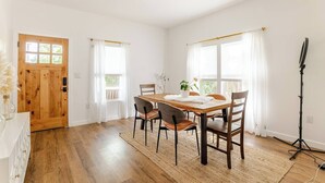 A dining area with a wooden table, four chairs, and a rug. There is a door with a window, a light fixture on the ceiling, and a microphone stand.