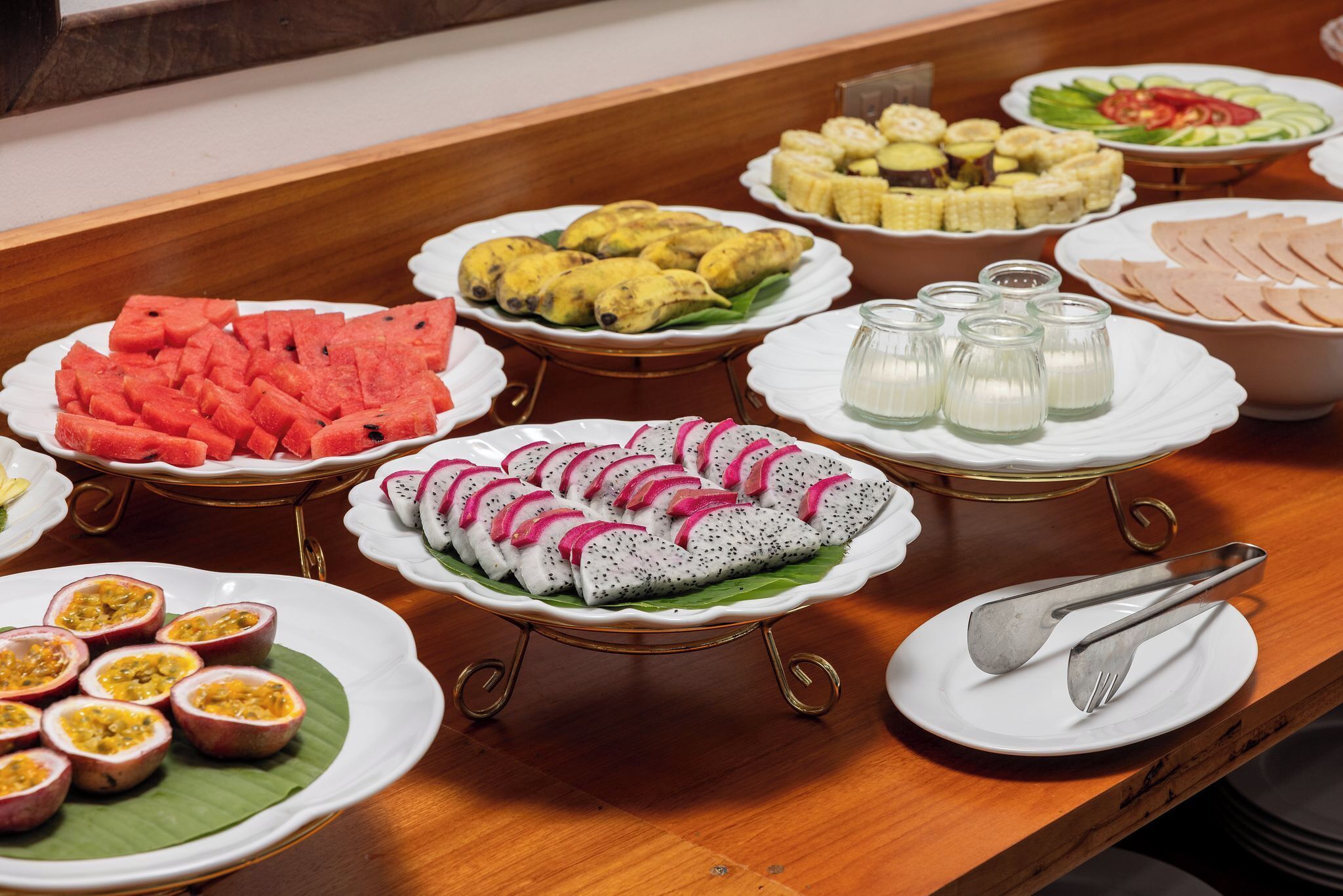 A buffet table with various dishes including watermelon, dragon fruit, and corn on the cob.