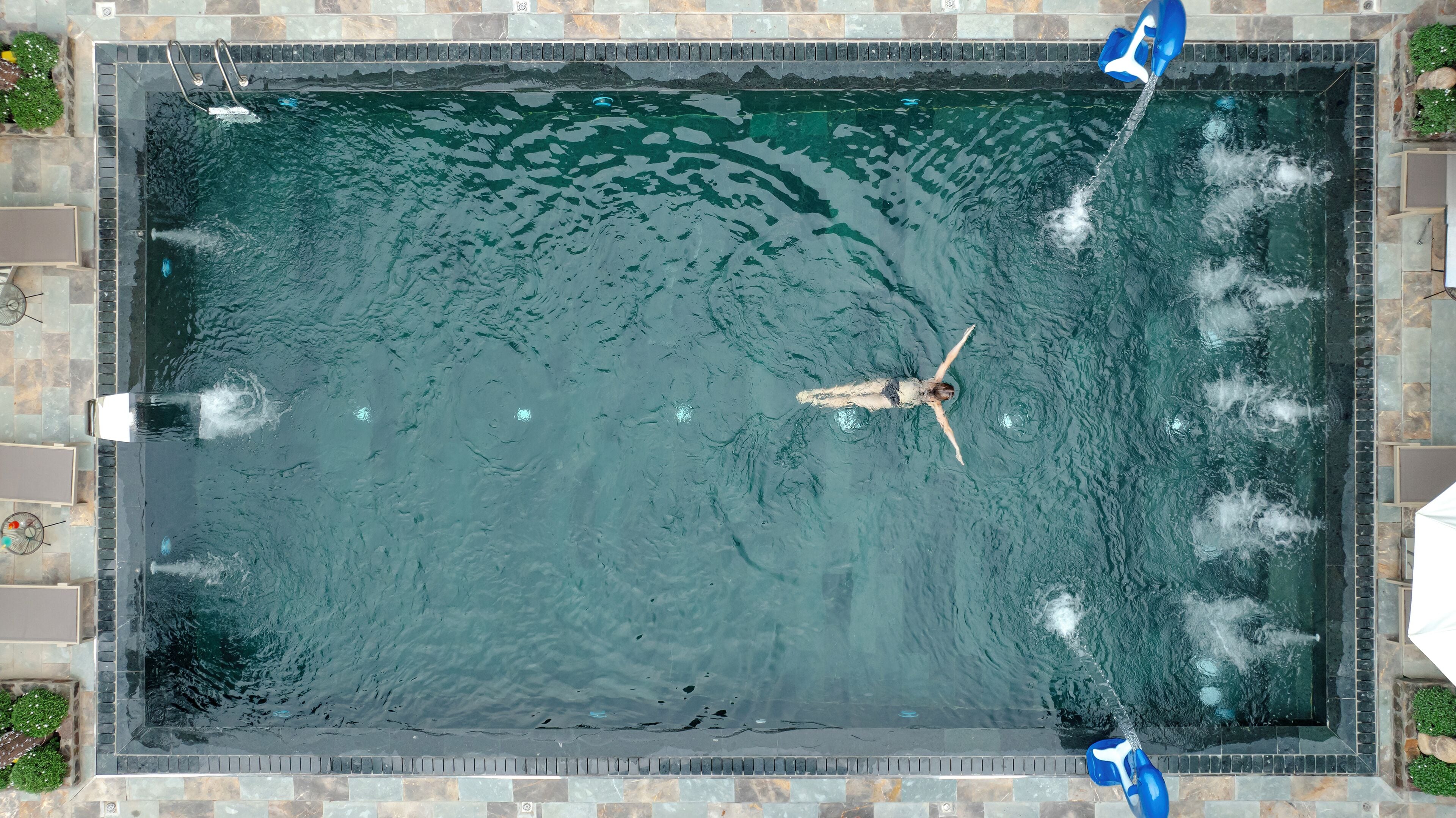 Vista aérea de una piscina rectangular con borde de piedra, rodeada por una zona de patio con mobiliario y plantas.