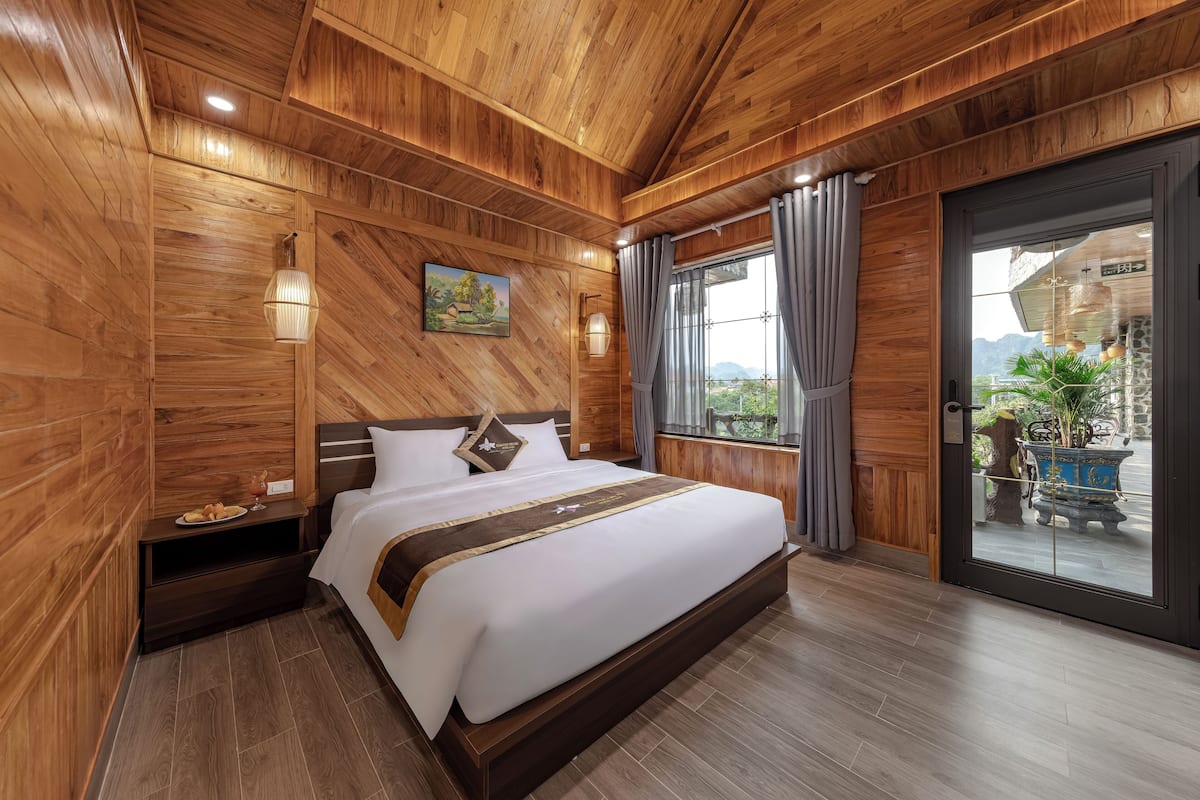 Une chambre à coucher aux murs lambrissés, avec un grand lit, une table de chevet, un tableau et une vue sur l’extérieur, visible à travers une porte vitrée.