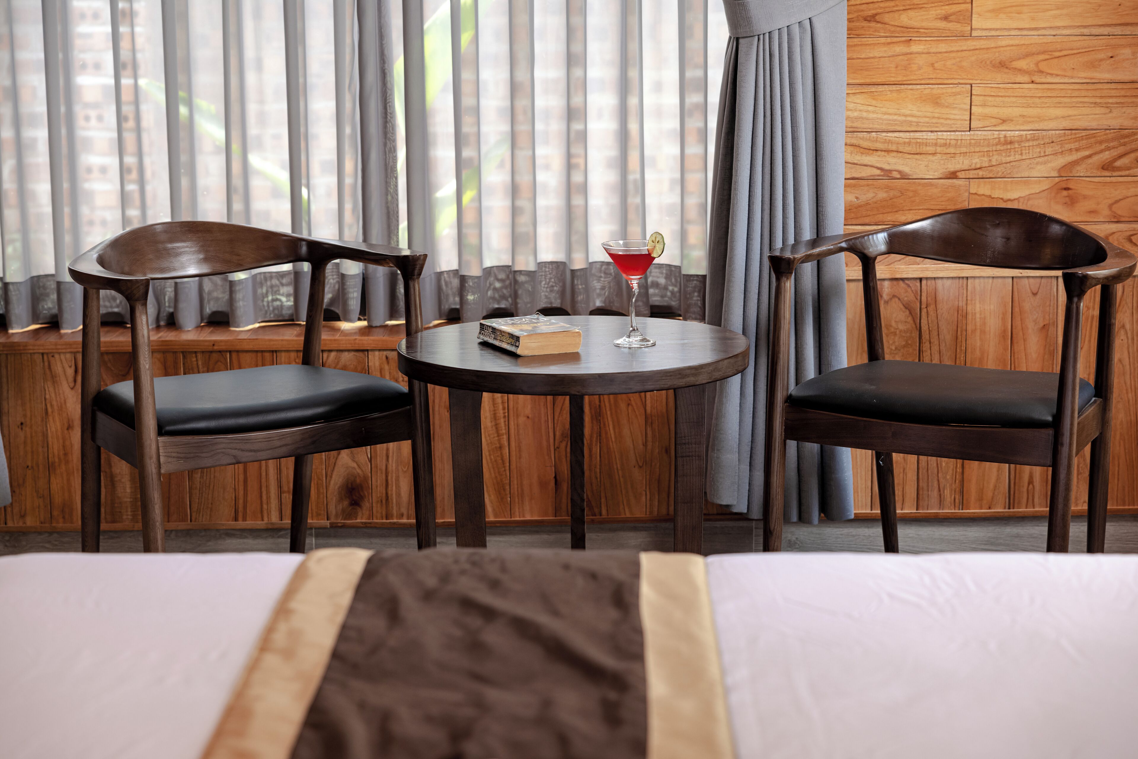 A hotel room with a bed, two wooden chairs, a small round table with a drink, and a book.