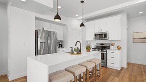 Une cuisine moderne avec des meubles blancs, des appareils en acier inoxydable, un îlot central doté de tabourets et un éclairage suspendu.