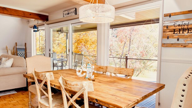 Un espace repas avec une table et des chaises en bois, un luminaire suspendu et une vue sur les arbres d’automne à travers de grandes fenêtres.