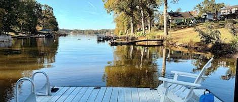 A wooden dock with a boat chair and a water cooler, overlooking a calm river with boats and trees.
