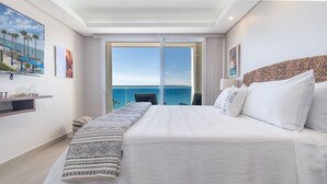 Een hotelkamer met een groot bed, uitzicht op de oceaan, een balkon met een stoel en een plank met decoratieve voorwerpen.
