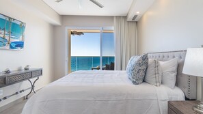 Une chambre d’hôtel avec un grand lit, une vue sur l’océan, un balcon avec une table et des chaises, et un tableau au mur.