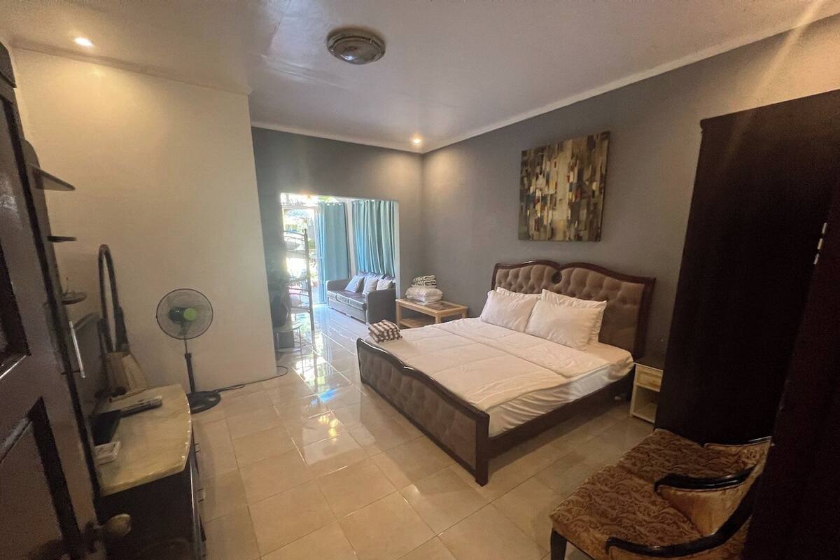 Une chambre d’hôtel avec un lit, une chaise, un bureau, un ventilateur et un tableau au mur.
