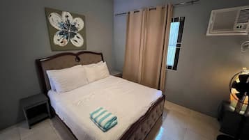 A neatly made bed with a striped towel, a small bedside table, a wall-mounted air conditioning unit, and a window with beige curtains.