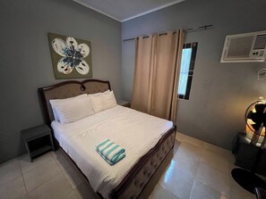 A neatly made bed with a striped towel, a small bedside table, a wall-mounted air conditioning unit, and a window with beige curtains. - Nora's Place Boutique Resort (Panglao)