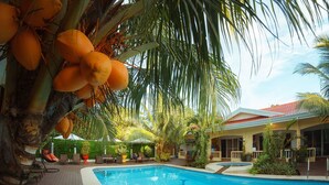 Une piscine dotée d’une terrasse carrelée, entourée de palmiers et bordée d’un bâtiment au toit rouge.