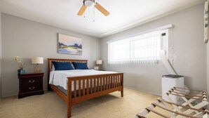 Une chambre à coucher avec un lit en bois, deux tables de chevet, un ventilateur de plafond, une fenêtre avec des stores et un radiateur.