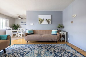 Un salon avec un canapé beige, un tapis à motifs bleus, un tableau représentant un sentier et un ventilateur de plafond.