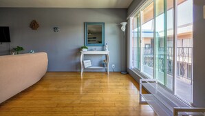 Une pièce avec un plancher en bois, un banc blanc, une petite table blanche avec un miroir et une grande fenêtre donnant sur l’extérieur.