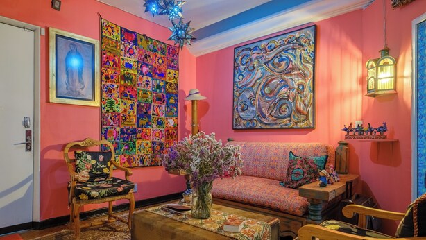 A cozy living room with a vibrant pink wall, a large colorful quilt, a sofa, a wooden coffee table with a vase of flowers, and a decorative lantern.