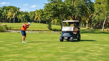 Un campo de golf con una persona, vestida con una camisa roja y pantalones cortos azules, preparándose para golpear una pelota, y un carrito de golf 'Club Car' estacionado sobre el césped.