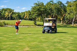 A golf course with a person in a red shirt and blue shorts preparing to hit a golf ball, and a golf cart labeled 'Club Car' parked on the grass. - High-End Waterfront Condo Secrets Waters (Puerto Aventuras)