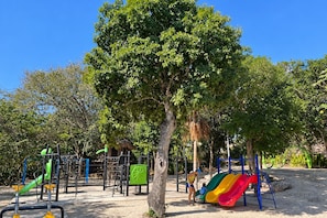A playground with various play structures including slides and climbing frames, surrounded by trees. - High-End Waterfront Condo Secrets Waters (Puerto Aventuras)