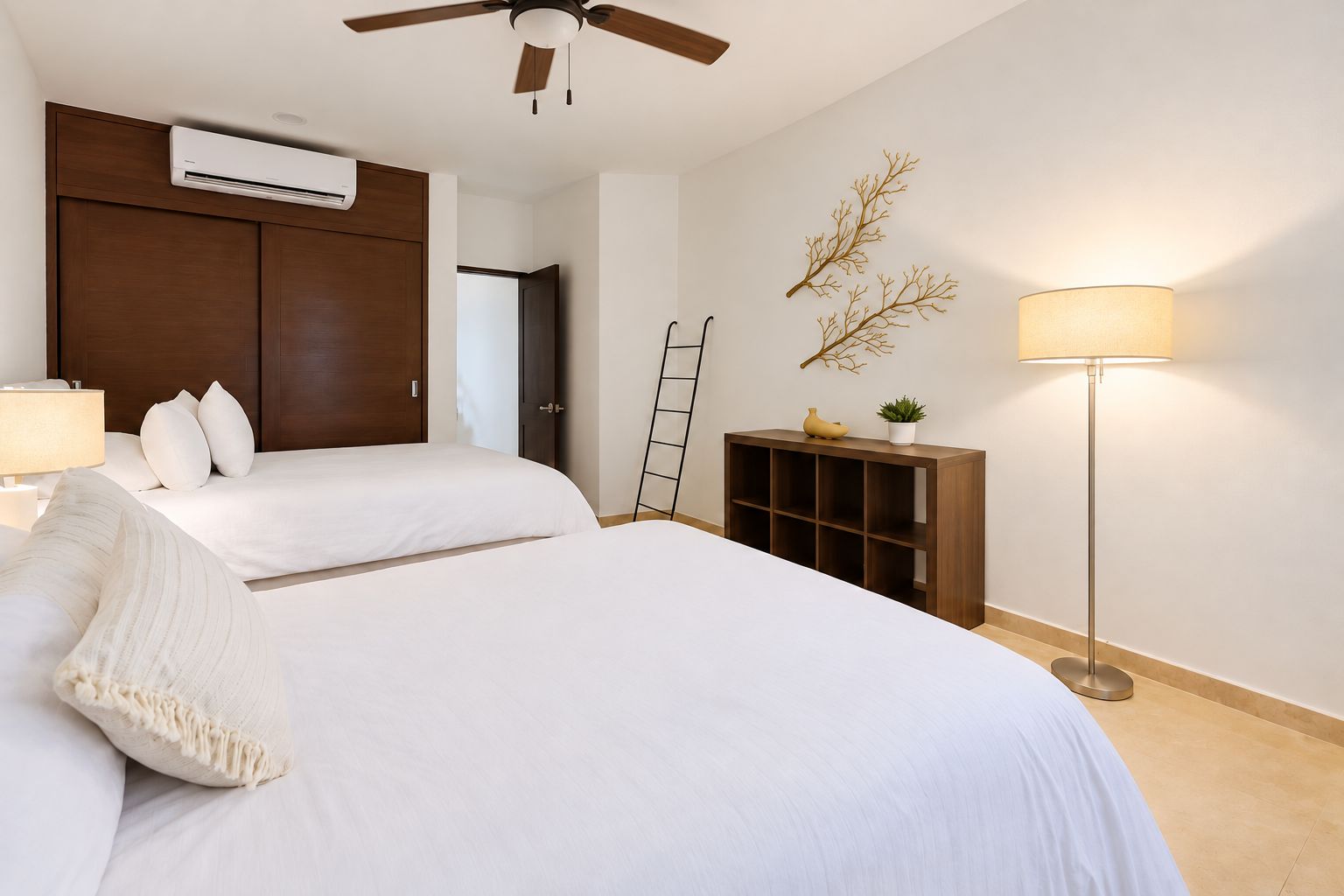 Habitación de hotel con dos camas, ventilador de techo, unidad de aire acondicionado, lámpara de noche, pequeña consola de madera y una rama decorativa en la pared.
