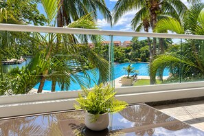 A balcony with a view of a swimming pool surrounded by palm trees and a clear sky. - High-End Waterfront Condo Secrets Waters (Puerto Aventuras)